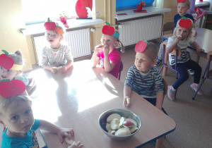 Dzieci siedzą przy stoliku i jedzą jabłka.