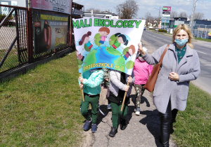 Dzieci z wychowawczynią niosą transparent "Mali Ekolodzy" i idą ulicami naszego miasta