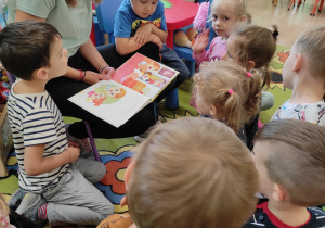 Dzieci siedzą na dywanie i słuchają opowieści czytanej przez mamę Olusia