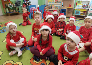 Dzieci siedzą na dywanie w czerwonych strojach i czapkach