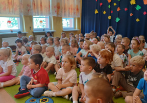 Dzieci siedzą na dywanie i oglądają występy muzyków