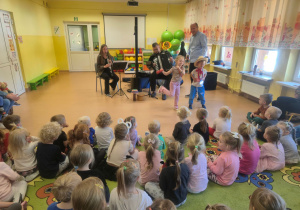 Dzieci siedzą na dywanie i oglądają występ muzyków
