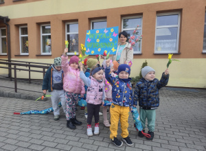 Dzieci stoją przed przedszkolem z transparentem "Witaj Wiosno"